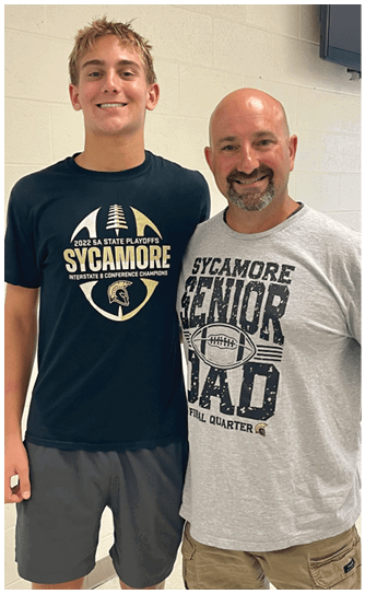 Two people smiling and standing close together indoors wearing Sycamore-themed t-shirts against a light-colored brick wall. Description generated by AI