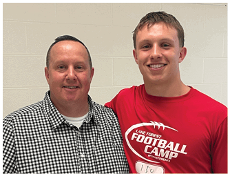 Two people stand close together with one wearing a black and white checkered shirt and the other wearing a red shirt that says “Lake Forest Football Camp" with a name tag. Description generated by AI
