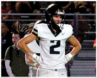 A person wearing a white football uniform with the number two stands with hands on hips on a field while people watch in the background. Description generated by AI