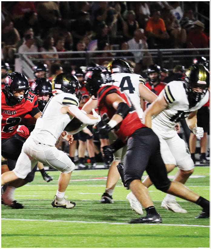 Players in red and black and players in white and black uniforms are actively engaged in a football game on a field with spectators in the background. Description generated by AI