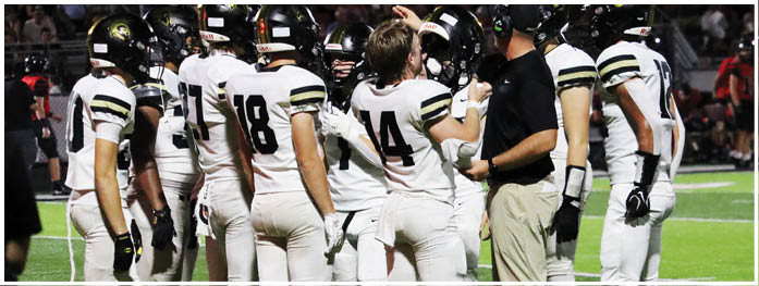 A group of people wearing white football uniforms with black and gold stripes are standing on a field while one person in a black shirt and khaki pants appears to be giving instructions. Description generated by AI