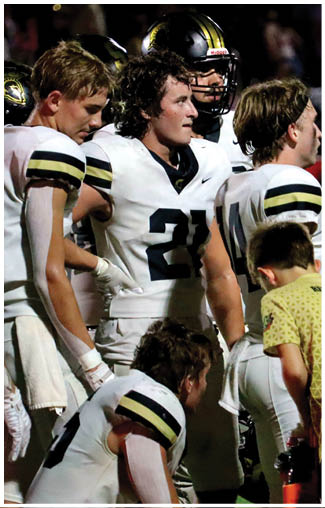 A child wearing a yellow shirt with text on the back is bending over near adults dressed in white football uniforms on a field. Description generated by AI