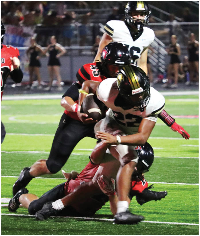 A person in a black and red football uniform tackles another person in a white and gold football uniform who is holding a football on a field with blurred spectators in the background. Description generated by AI