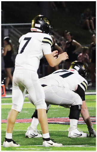 Two people in white football uniforms and black helmets are positioned on a football field, with one standing behind the other who is crouched down. Description generated by AI