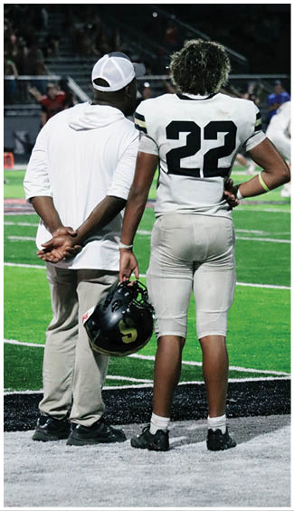 A person wearing a white sports jersey with the number 22 stands next to another person in a white shirt on a sports field at night. Description generated by AI