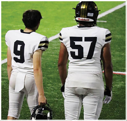 Two people wearing white football uniforms with numbers nine and fifty-seven stand on a green field, with one holding a helmet. Description generated by AI