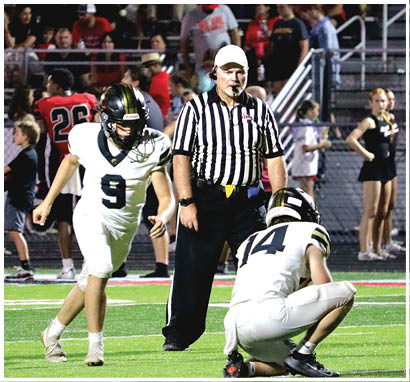 A referee stands on a football field between two players in white uniforms with black helmets while a crowd watches from the bleachers in the background. Description generated by AI