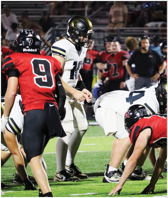 Several people in football uniforms are positioned on a field, with one person in a red jersey numbered 9 standing behind others in white and black uniforms preparing for a play. Description generated by AI