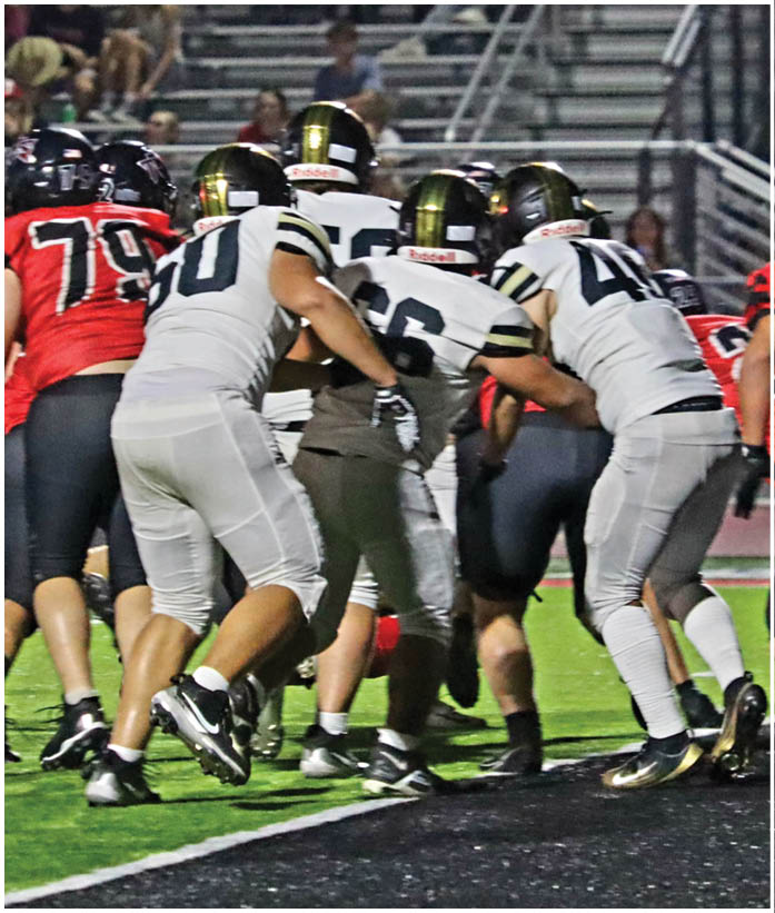 Several people in football uniforms are engaged in a play on a field with artificial turf and stadium seating in the background. Description generated by AI