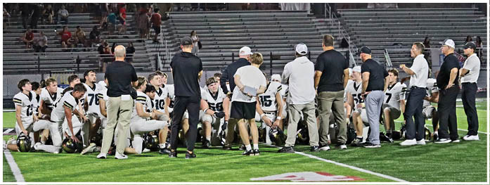 A group of people in sports uniforms and coaches in casual clothing are gathered on a football field, with some sitting and others standing, likely during a team meeting or timeout. Description generated by AI