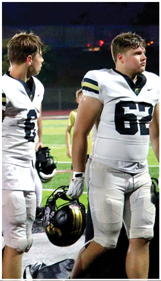A person in a white football uniform with black and gold stripes is standing on a field holding two black helmets while looking to the side. Description generated by AI