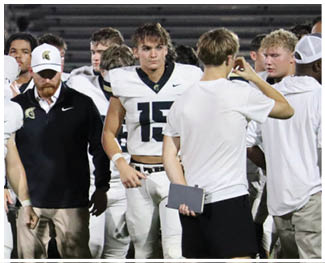 A group of people in football uniforms and coaching attire stand together on a field with bleachers in the background. Description generated by AI