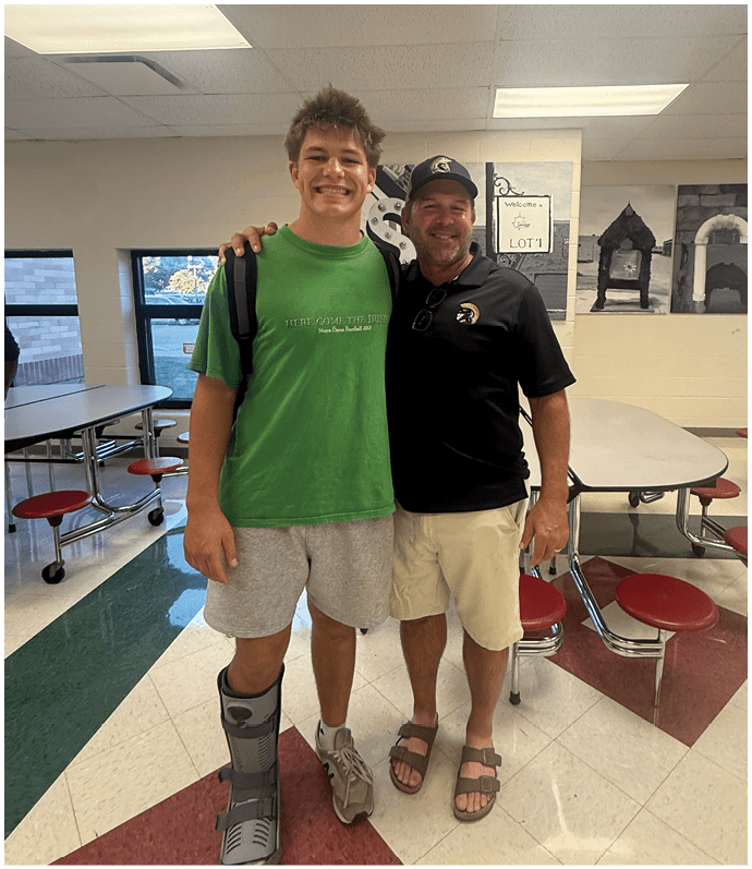 Two people stand closely together smiling in a cafeteria with red stools and tables, one wearing a green shirt and gray shorts with a leg brace, and the other wearing a black shirt, beige shorts, and a hat. Description generated by AI