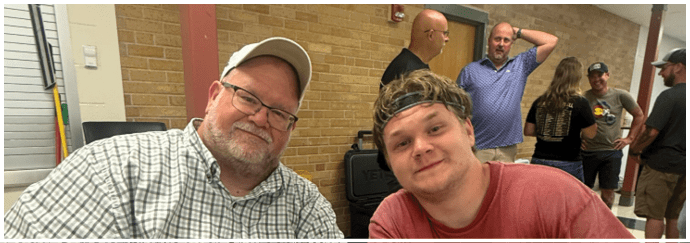 Two adults sitting and smiling at a table in a casual indoor setting with several other people standing and talking in the background. Description generated by AI