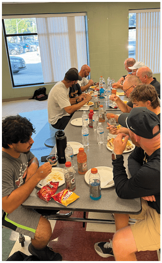 Several people sit closely together at a long table in a casual indoor setting, eating and drinking while engaging in conversation. Description generated by AI