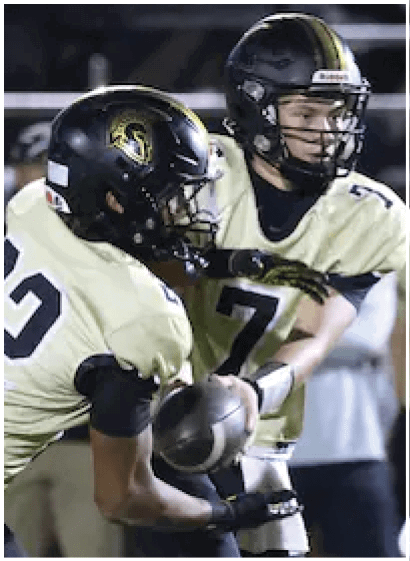 Two people wearing football helmets and uniforms are engaged in a play with one person handing off the football to the other on a field. Description generated by AI