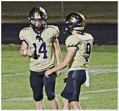 Two people wearing football uniforms with helmets are standing on a grassy field, with one giving a thumbs-up gesture. Description generated by AI