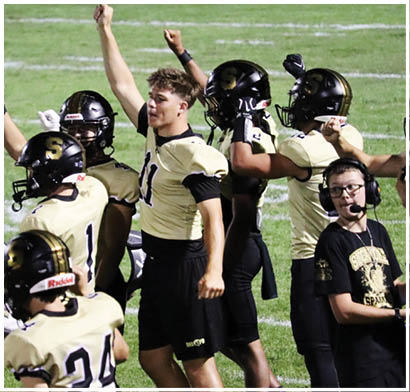 A group of people wearing football uniforms and helmets are celebrating on a field while another person with glasses and a headset stands nearby. Description generated by AI
