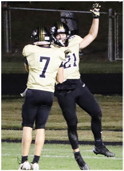Two people wearing football uniforms with helmets are celebrating on a field, one jumping with an arm raised and the other standing with the number 7 visible on their jersey. Description generated by AI