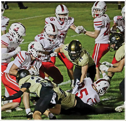 Several people wearing football helmets and uniforms are engaged in a play on a grassy field, with one person in a gold and black uniform bending forward while others in white and red uniforms surround them. Description generated by AI