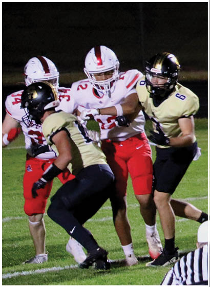 Four people in football uniforms are engaged in a play on a grassy field at night with one person wearing a striped referee shirt partially visible in the foreground. Description generated by AI
