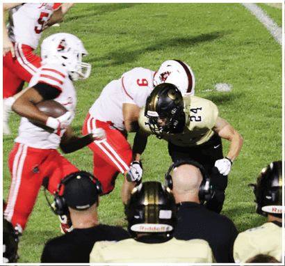 Two football players in white and red uniforms are running on a field while a player in a black and gold uniform is crouched down near the sideline with coaches watching. Description generated by AI
