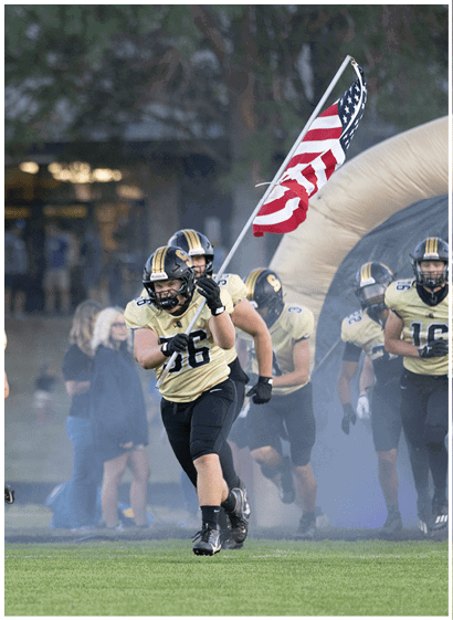 A person wearing a football uniform and helmet is running while holding an American flag with other players and people in the background. Description generated by AI