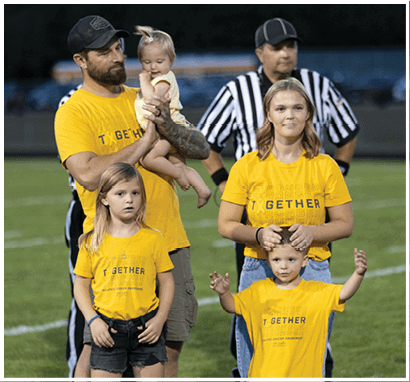 A group of adults and children wearing matching yellow shirts stand together on a sports field with a referee in the background. Description generated by AI