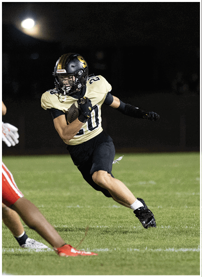 A person wearing a black and gold football uniform is running on a grassy field at night while another person in red pants is partially visible. Description generated by AI