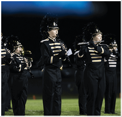 A person in a black and gold marching band uniform with a tall feathered hat is playing a clarinet during a nighttime performance. Description generated by AI