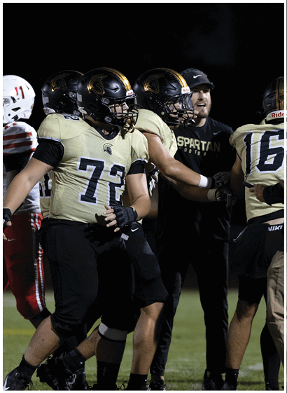 Several people wearing football uniforms and helmets are gathered on a field at night, with one person in a black shirt and cap interacting with the players. Description generated by AI