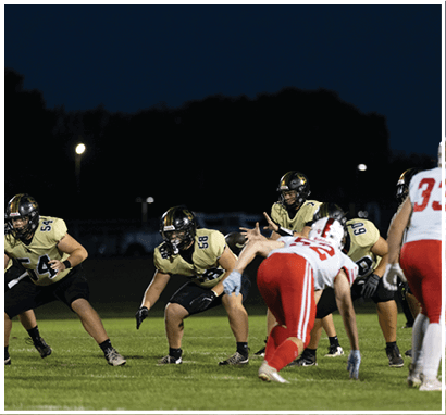 People wearing football uniforms are positioned on a field at night, preparing for a play with some in a crouched stance and others standing. Description generated by AI