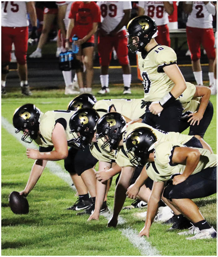 Several people wearing black helmets and light-colored sports uniforms are crouched in a line on a grassy field, appearing to prepare for a play in a football game. Description generated by AI