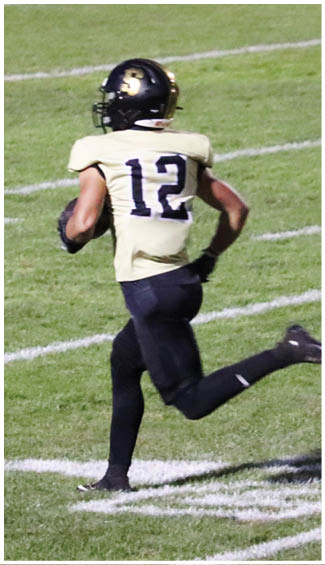 A person wearing a black helmet and a beige jersey with the number 12 is running on a grassy field while holding a football. Description generated by AI