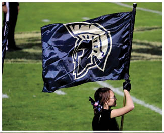 A person is holding a dark blue flag with a white Spartan helmet logo on a grassy field. Description generated by AI
