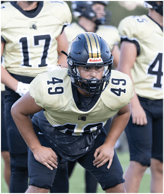A person wearing a football helmet and uniform with the number 49 is in a ready stance on a field with other players in similar uniforms in the background. Description generated by AI