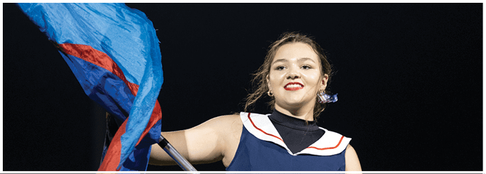 A person wearing a navy blue outfit with a white and red collar is holding and waving a blue and red flag against a dark background. Description generated by AI