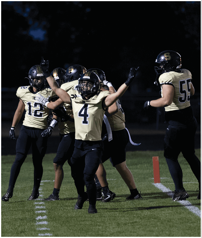 Five people in football uniforms are on a field at night with one person in the center raising their arms in celebration. Description generated by AI