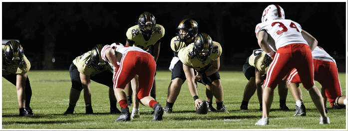 People in football uniforms are positioned on a field at night, preparing for a play. Description generated by AI