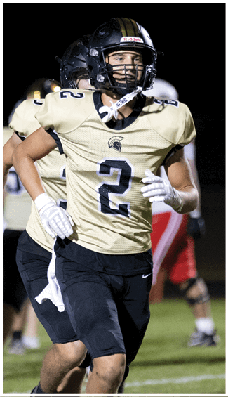 Two people wearing football uniforms and helmets are running on a field during a nighttime game. Description generated by AI