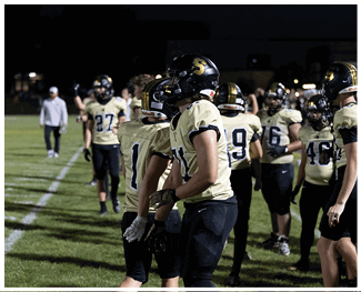 People wearing football uniforms and helmets stand on a grassy field, some with gloves, appearing to prepare for or participate in a game. Description generated by AI