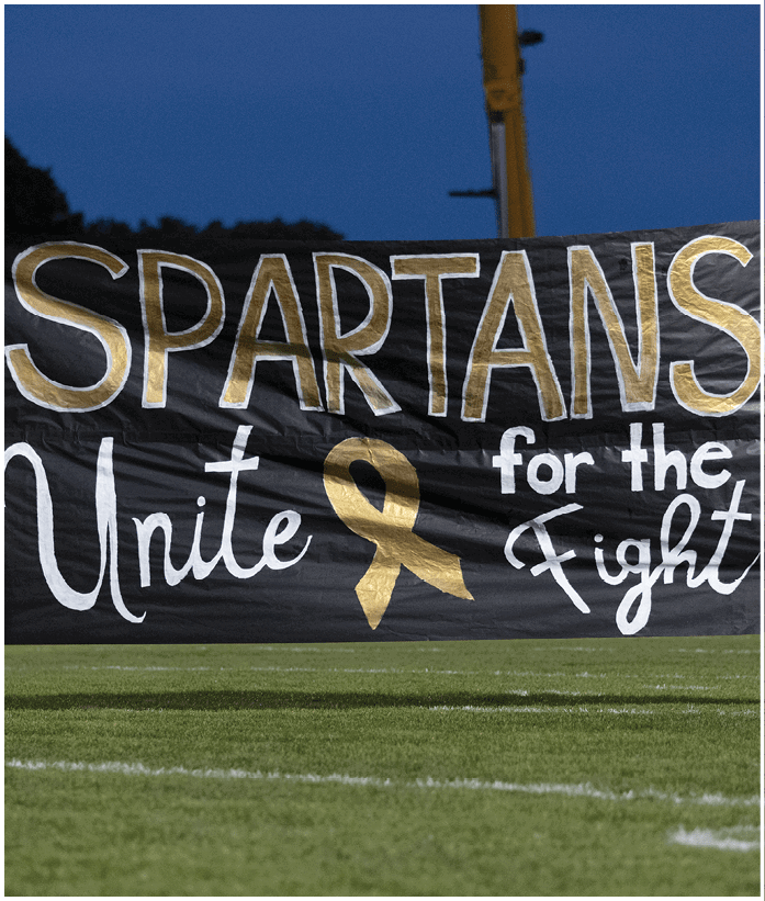 People holding yellow pom-poms stand next to a large black banner on a grassy field at dusk that reads “SPARTANS Unite" with a gold ribbon symbol. Description generated by AI