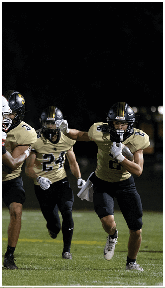 Three people in football uniforms are actively playing on a field at night, with one person running while holding the ball and another person attempting to block an opponent. Description generated by AI