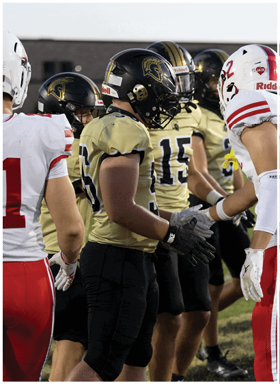 People wearing football uniforms from two different teams are standing on a field, appearing to prepare for or discuss the game. Description generated by AI