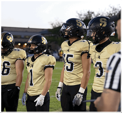 Four people wearing football uniforms and helmets stand on a grassy field during what appears to be a game or practice at dusk. Description generated by AI