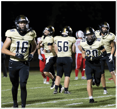 Several people wearing football uniforms with helmets are on a grassy field at night, with some in light-colored jerseys and others in red and white jerseys in the background. Description generated by AI