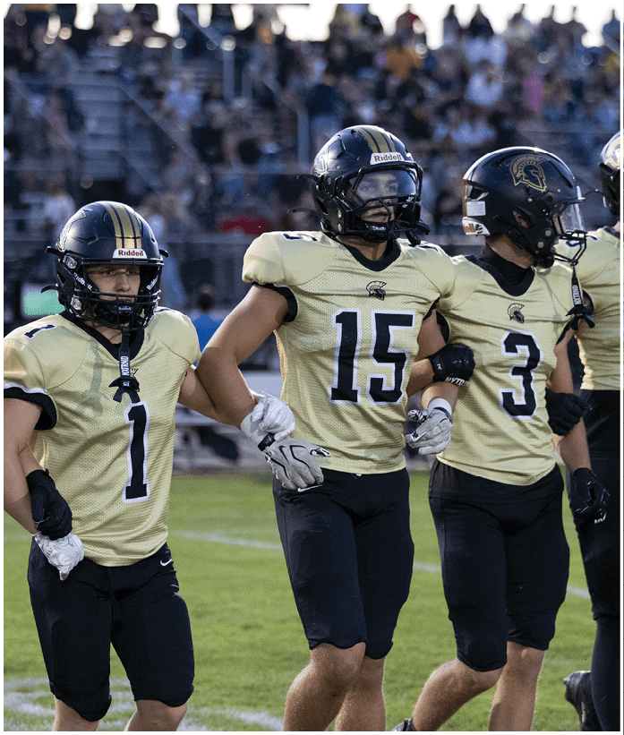 A person wearing a football helmet and uniform with the number one is standing on a field holding hands with two other people also in football uniforms. Description generated by AI