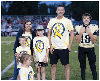 A group of people wearing matching yellow tie-dye shirts with a large yellow ribbon and the words Pediatric Cancer Awareness stand together on a sports field. Description generated by AI