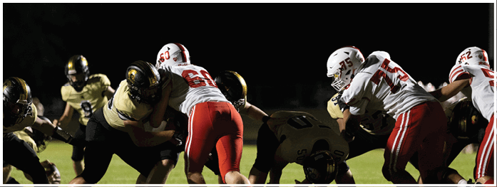 People in football uniforms are engaged in a close physical play on a field under stadium lighting. Description generated by AI