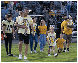 A group of people wearing matching yellow and white shirts with a ribbon design are walking on a grassy field in front of bleachers with spectators. Description generated by AI